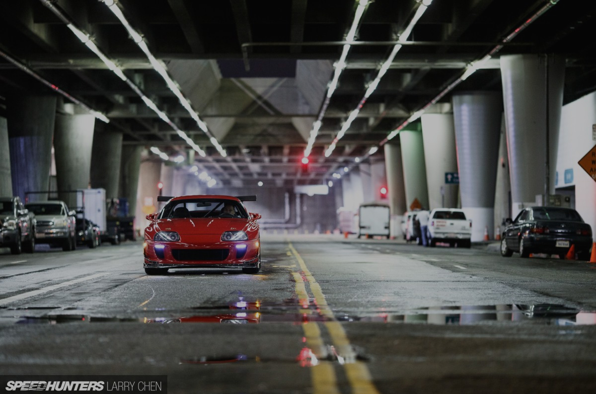 A red performance car photographed beneath city overpasses on a wet street